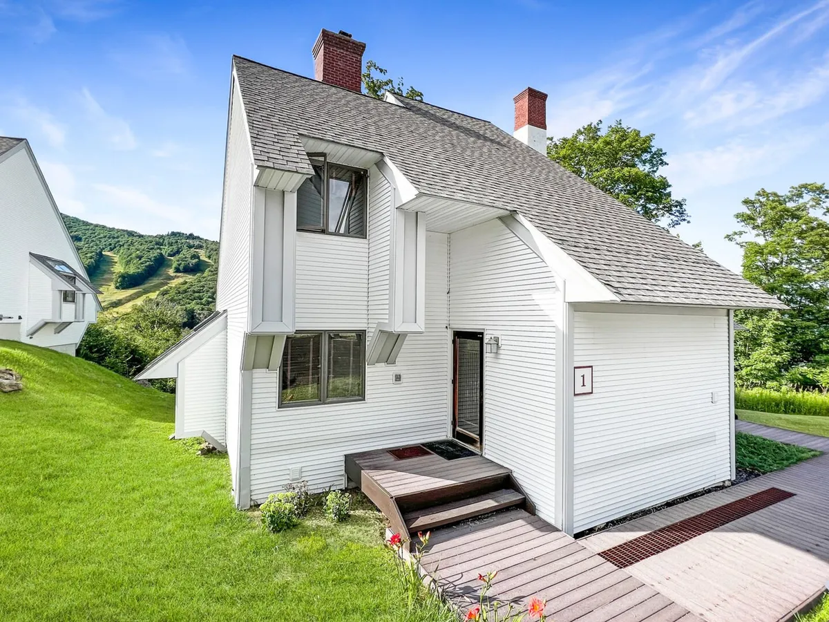 A view of the home's modern exterior architecture with a landscaped lawn, steps leading to the entrance, and surrounding hills. The scene is inviting and suggests arrival, homecoming, and connection to nature.