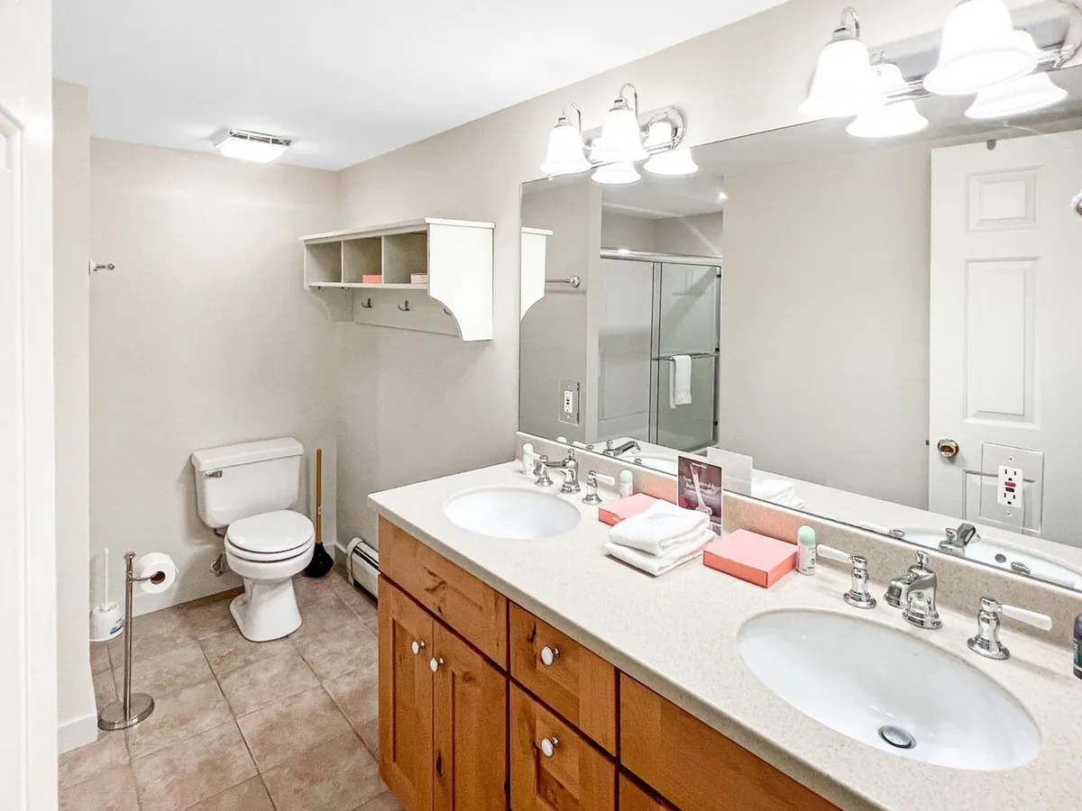 A spacious, spa-inspired bathroom featuring a double sink vanity, a large mirror, an inviting shower area, and a storage shelf. The clean lines and soothing lighting create an environment ideal for self-care and relaxation after a day outdoors.