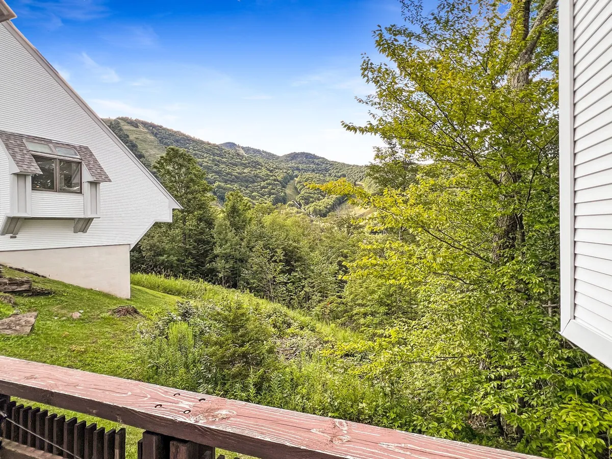 A wide view from the deck overlooking lush greenery and majestic mountains in the distance, framed by a classic deck railing. The scene evokes serenity and connection to the outdoors, with ample natural light and a hint of the home's inviting exterior atmosphere.