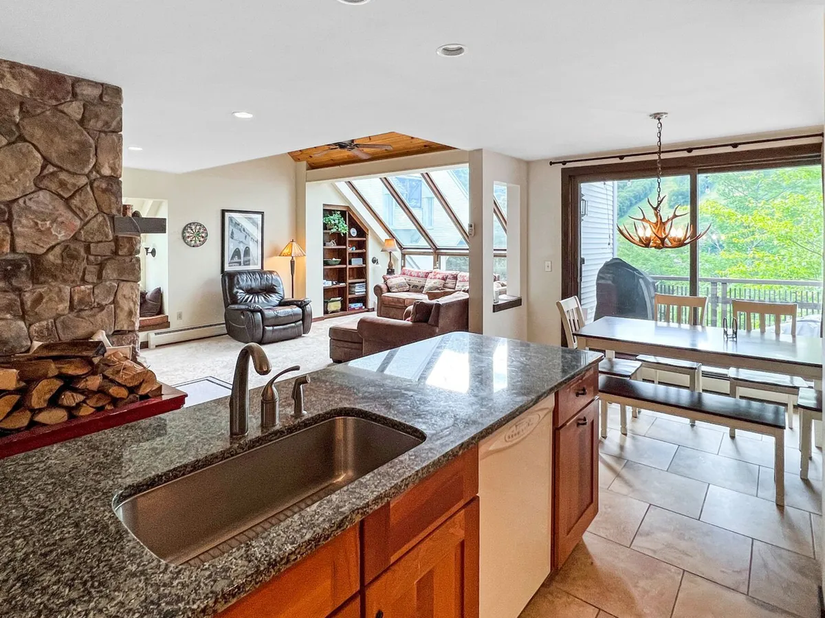 A welcoming great room combining a granite-topped kitchen, a large wooden dining table, a stone fireplace, and expansive windows revealing mountain vistas. The space is designed for both culinary activity and relaxation by the fire, with a seamless transition from kitchen to living and dining zones.