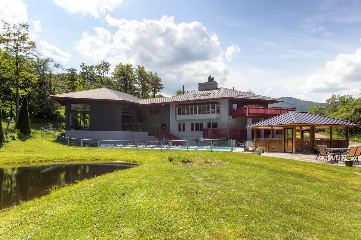 A wide exterior shot of the property highlighting its modern architecture, expansive lawn, outdoor seating area, and a tranquil pond, all set against lush greenery. The mountain horizon and open sky in the background evoke a peaceful, immersive experience in nature.