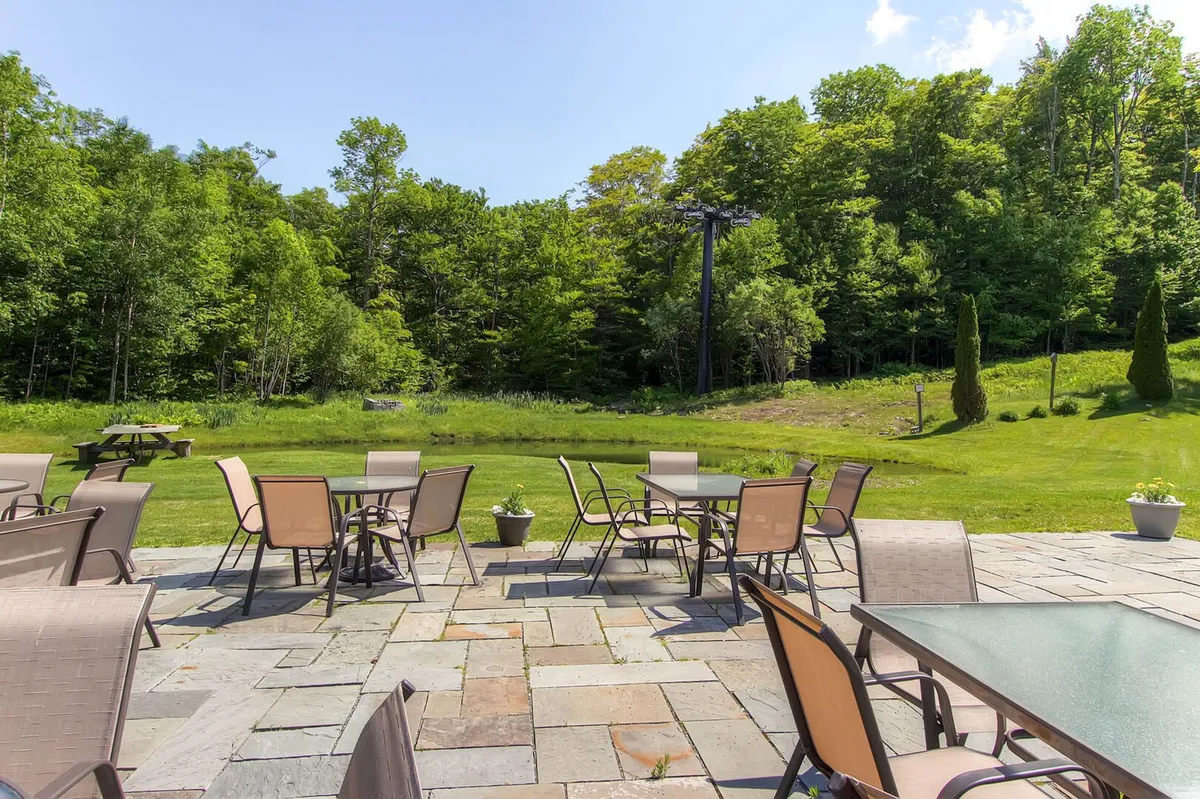 A tranquil stone patio with comfortable outdoor seating surrounded by lush greenery and a dramatic mountain backdrop. The space is set for outdoor dining or socializing, and the natural setting emphasizes relaxation and communal experiences.
