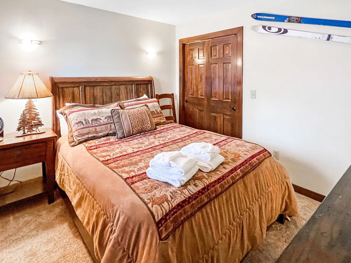 This image features a queen bedroom with a cozy bed dressed in decorative pillows, a nightstand with a warm lamp, a wooden closet door, and skis mounted on the wall. The space is inviting, filled with soft light, and radiates comfort—ideal for relaxation after a day’s activities.