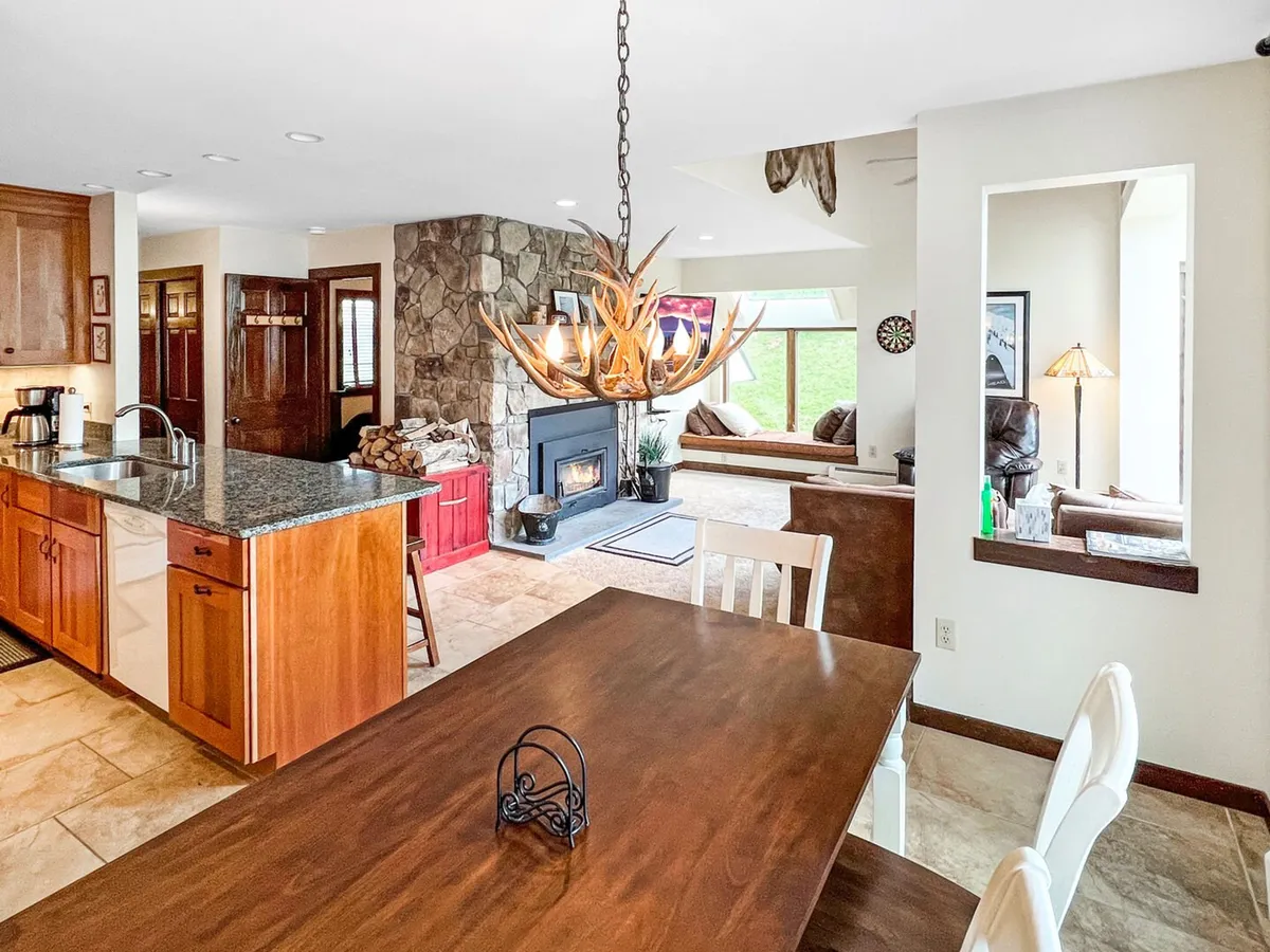 An inviting interior where a central kitchen island overlooks a cozy dining table and a stone fireplace. The open design ensures the chef is always part of the action, while the chandelier and natural materials add a touch of rustic elegance, making the space ideal for family meals and casual gatherings.