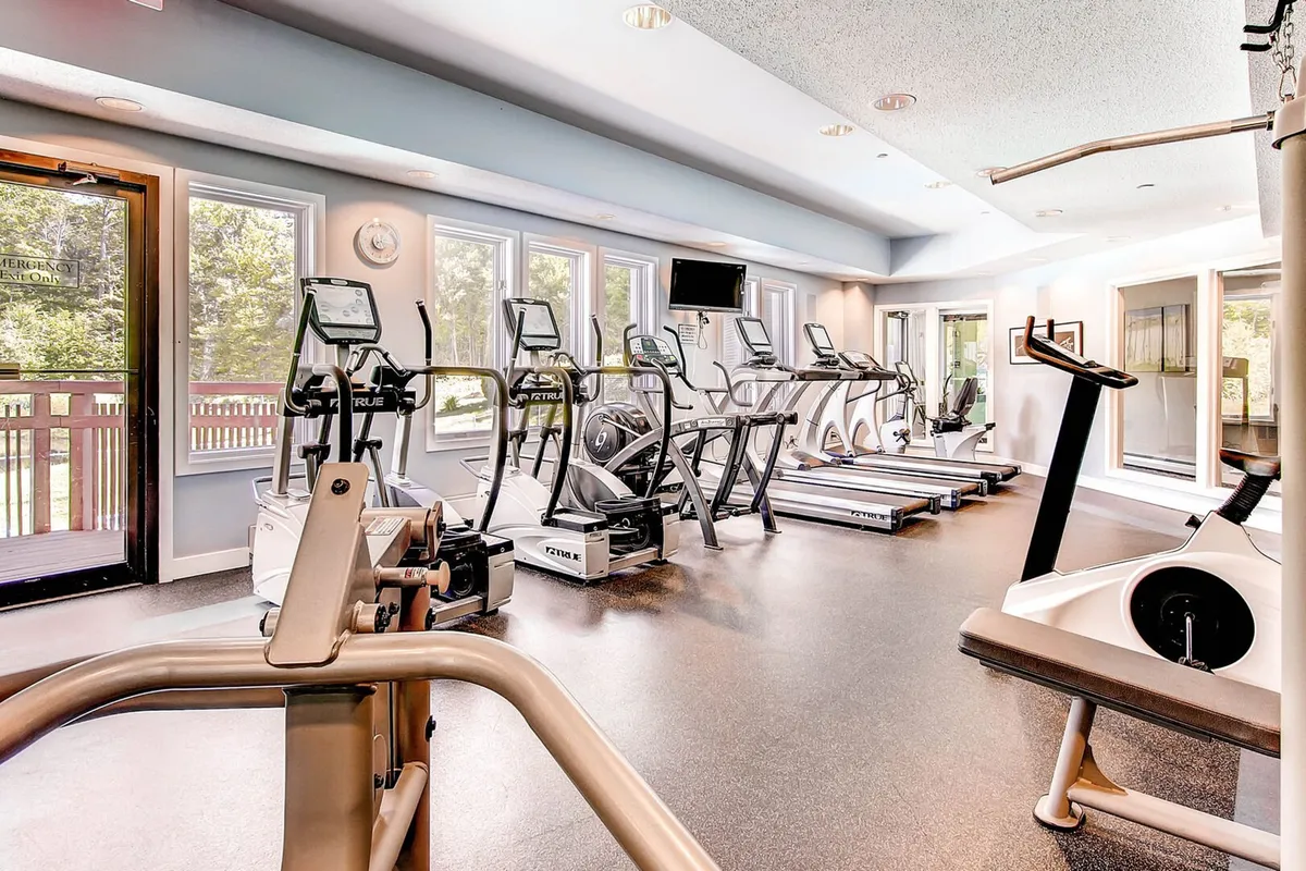 A modern, well-equipped fitness center with elliptical machines, treadmills, and a television, all facing large windows for uplifting natural light. The contemporary setup provides a motivating environment for maintaining fitness during your stay.