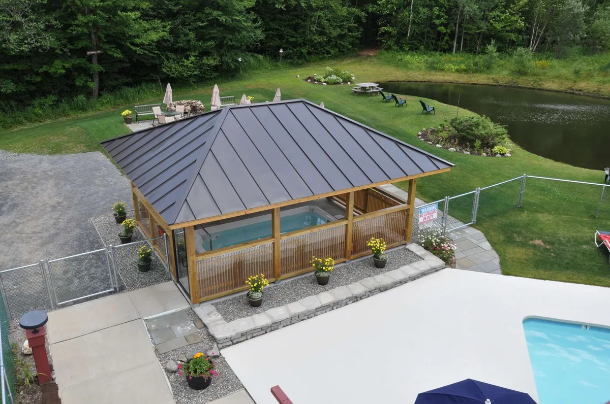 A tranquil exterior scene featuring a gazebo with a hot tub, lush landscaping, a pond in the background, and a pool area. The area is set up for relaxation and social activities, with people lounging and enjoying the space.