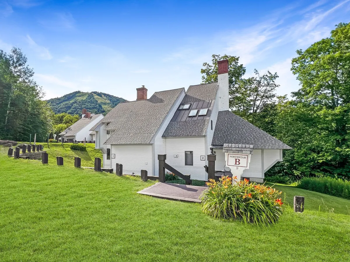 A view of the townhouse's exterior, showcasing the house structure nestled within a landscaped garden with a scenic mountain backdrop. The setting emphasizes proximity to nature and the outdoors.