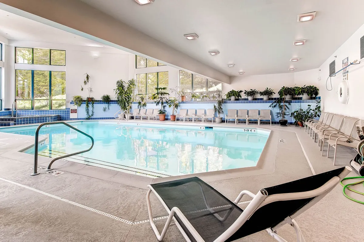 An inviting indoor swimming pool area with expansive lounge chairs, large windows that let in natural light, and lush indoor plants. The space exudes a tranquil, rejuvenating spa-like atmosphere perfect for relaxation and year-round enjoyment.