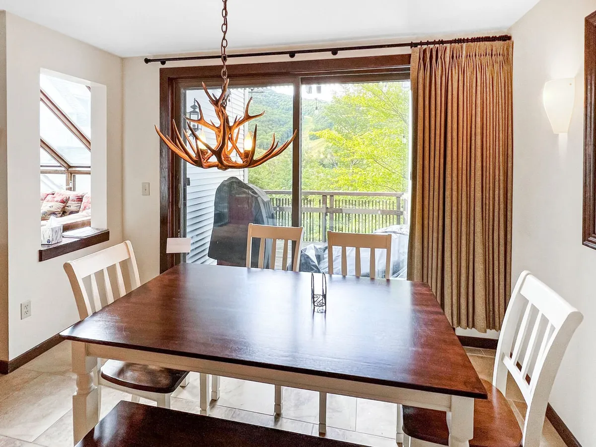 A cozy dining area featuring a sturdy wooden table and an antler chandelier, with a large glass door that opens to the deck and views of the outdoors. The space is designed for both intimate family dinners and lively gatherings, seamlessly connecting indoor and outdoor living.