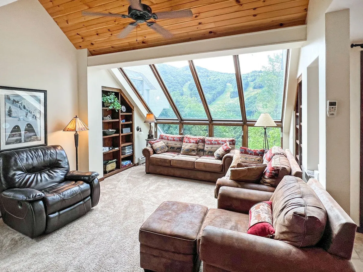 The image showcases a sunlit living area with expansive windows framing breathtaking mountain views, a plush seating area perfect for relaxation, a warm wooden ceiling, a stylish lamp, and a bookshelf. The scene feels inviting and tranquil, ideal for cozy evenings or gathering with loved ones.