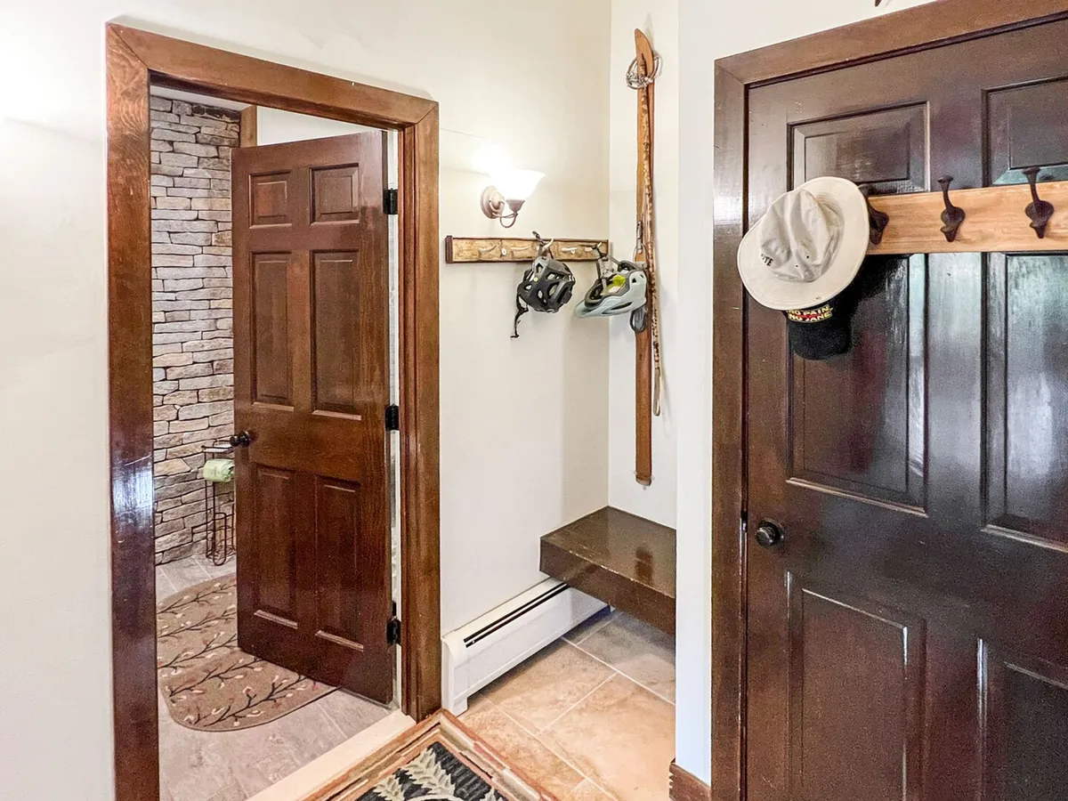 A functional, welcoming entryway featuring wooden doors, a stone wall, and a hat rack stocked with coats and helmets. The space is designed for easy access and storage of outdoor adventure gear, suggesting guests regularly come and go for skiing, hiking, or other activities.
